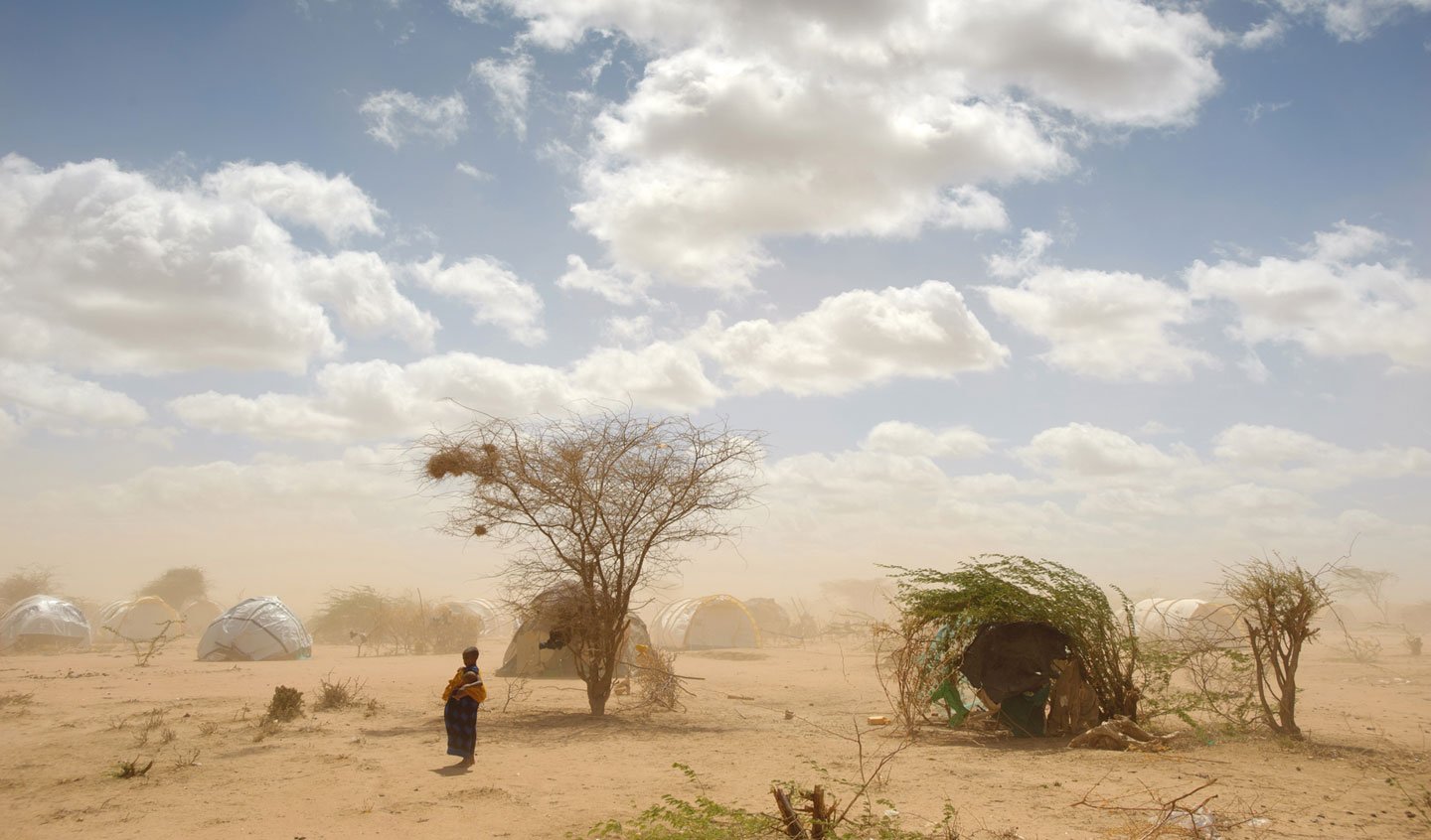 In Dabaad, a dusty refugee camp in Kenya, an African woman stands surrounded by makeshift shelters.