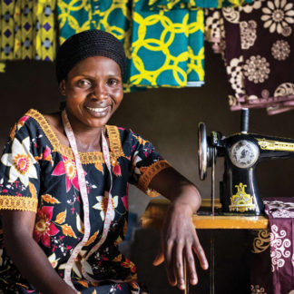 Woman smiles with her sewing machine with colorful fabrics hanging behind her.