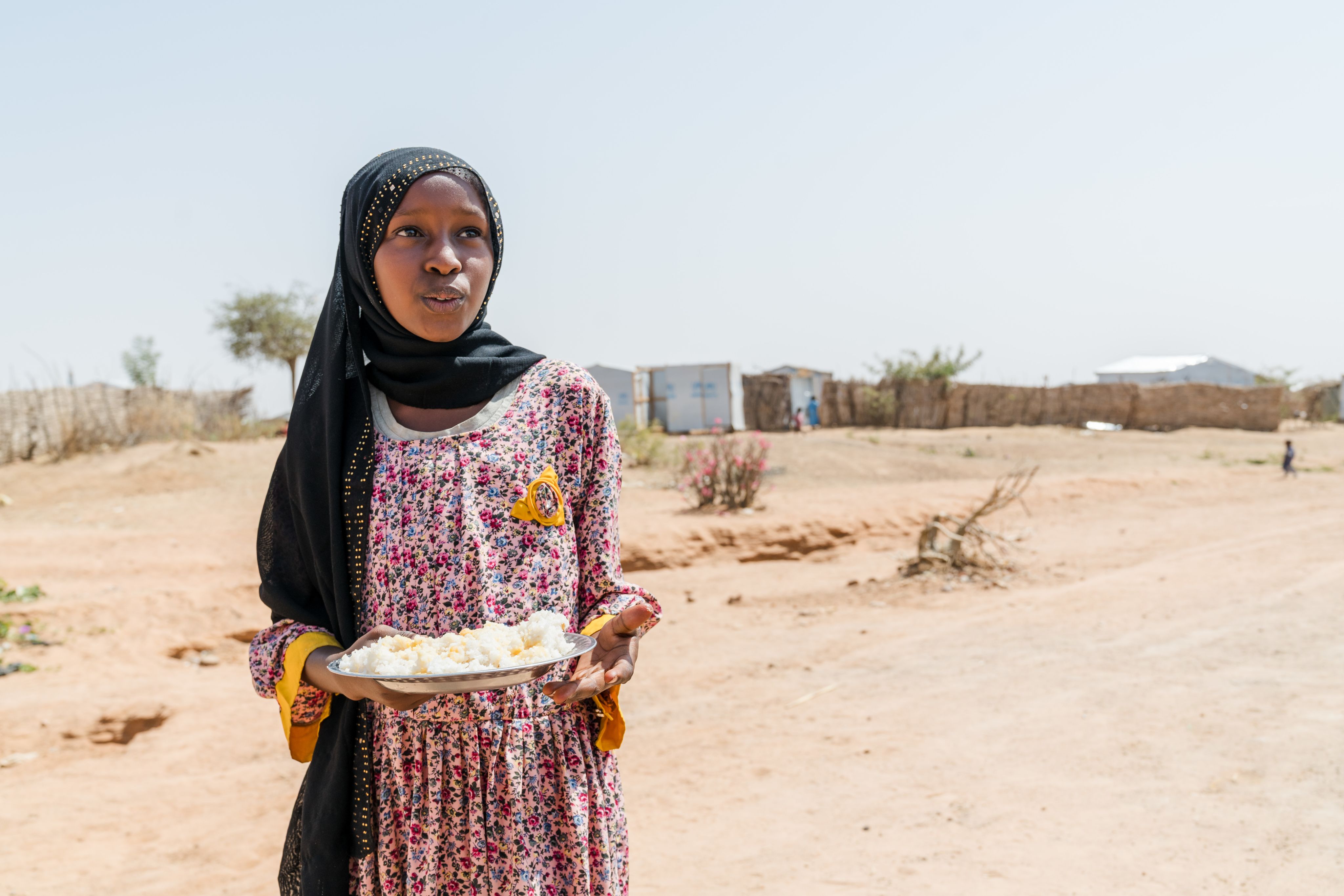 A teen girl holds a plate of food as she walks away from distant tents and looks into the distance under the midday sun.