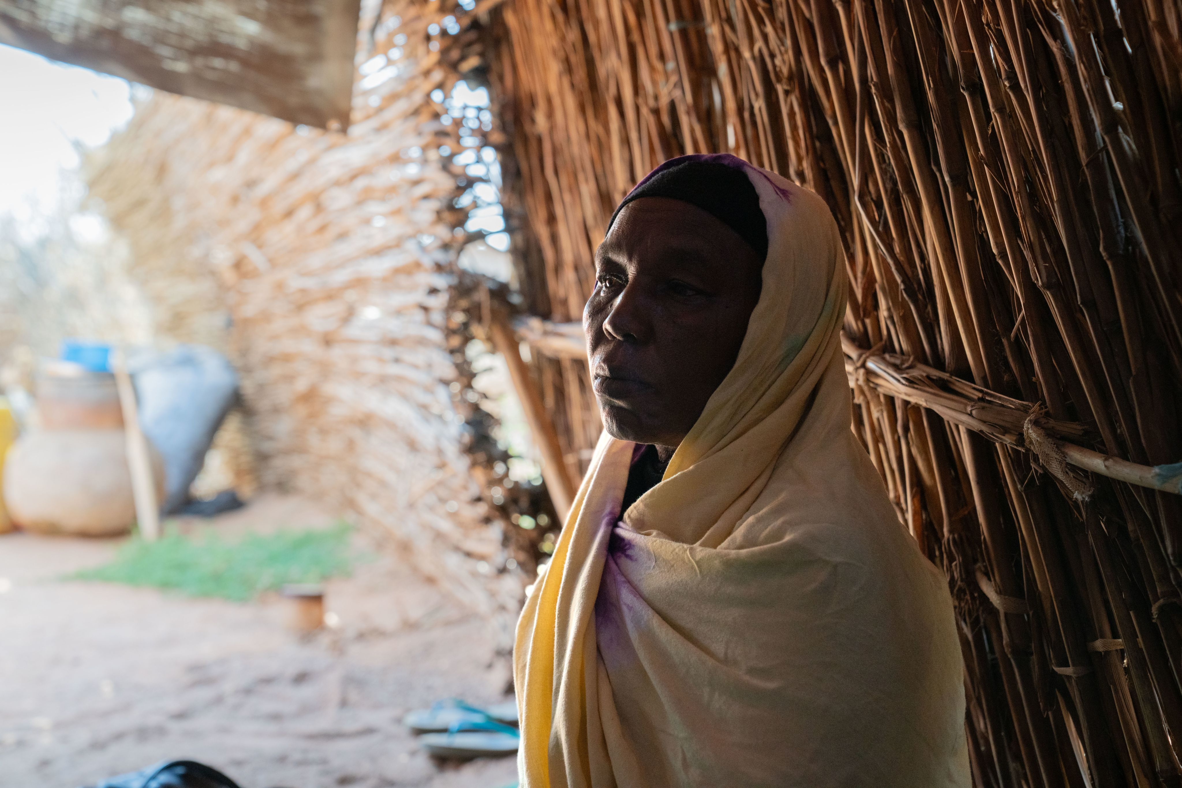 A woman in a yellow shawl sits quietly inside a shelter made of woven reeds, with soft light filtering through.