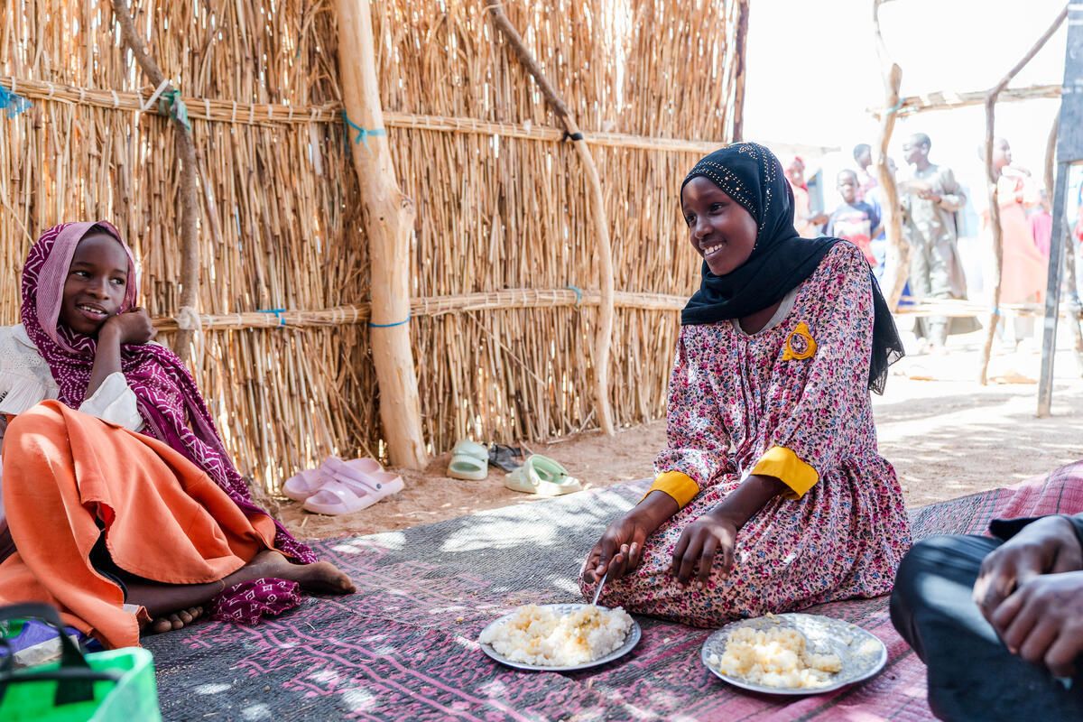 Two girls sit on a mat inside a thatch-walled shelter, smiling and eating plates of food.