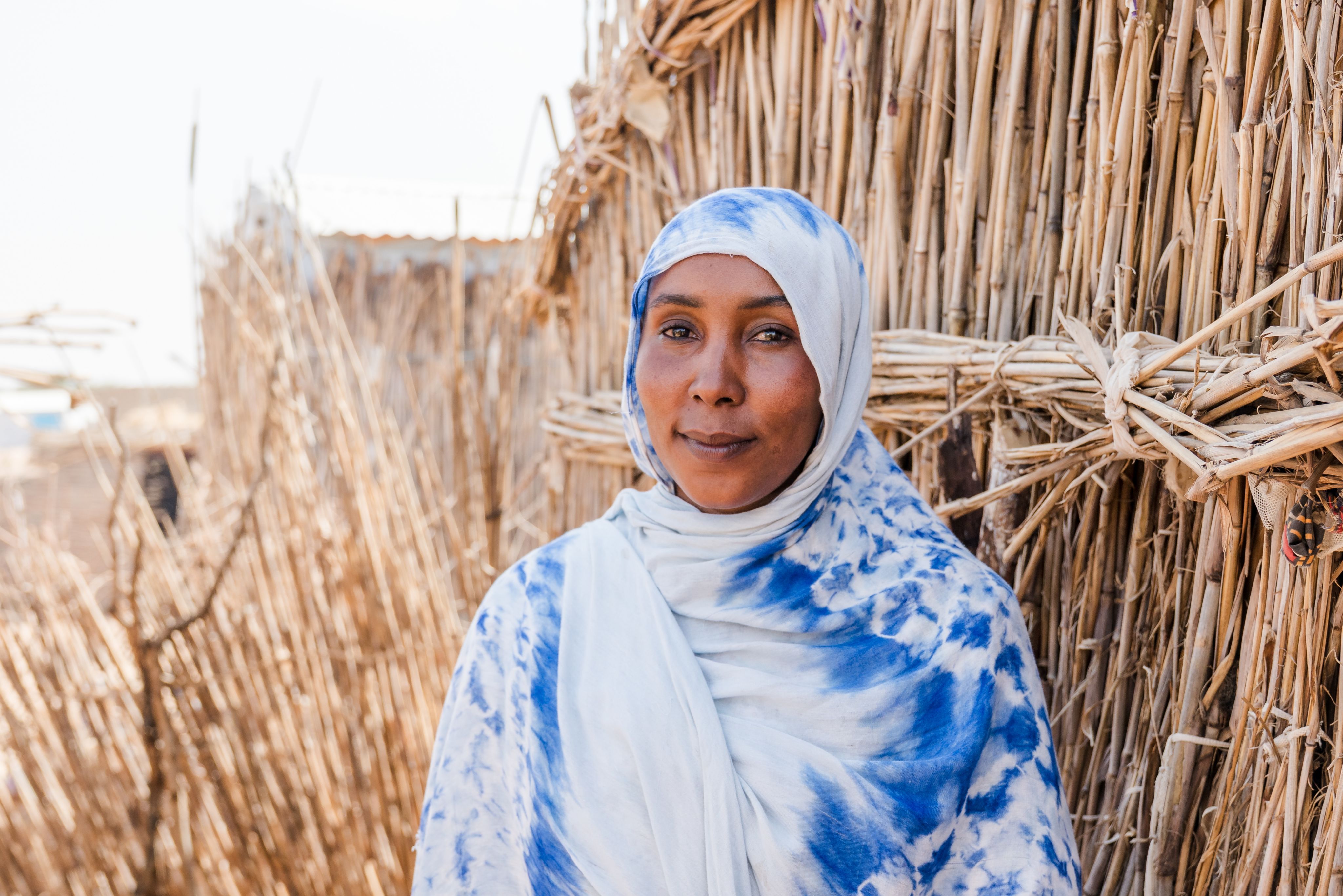 A woman in a blue and white headscarf stands beside a woven reed wall, looking calmly at the camera.