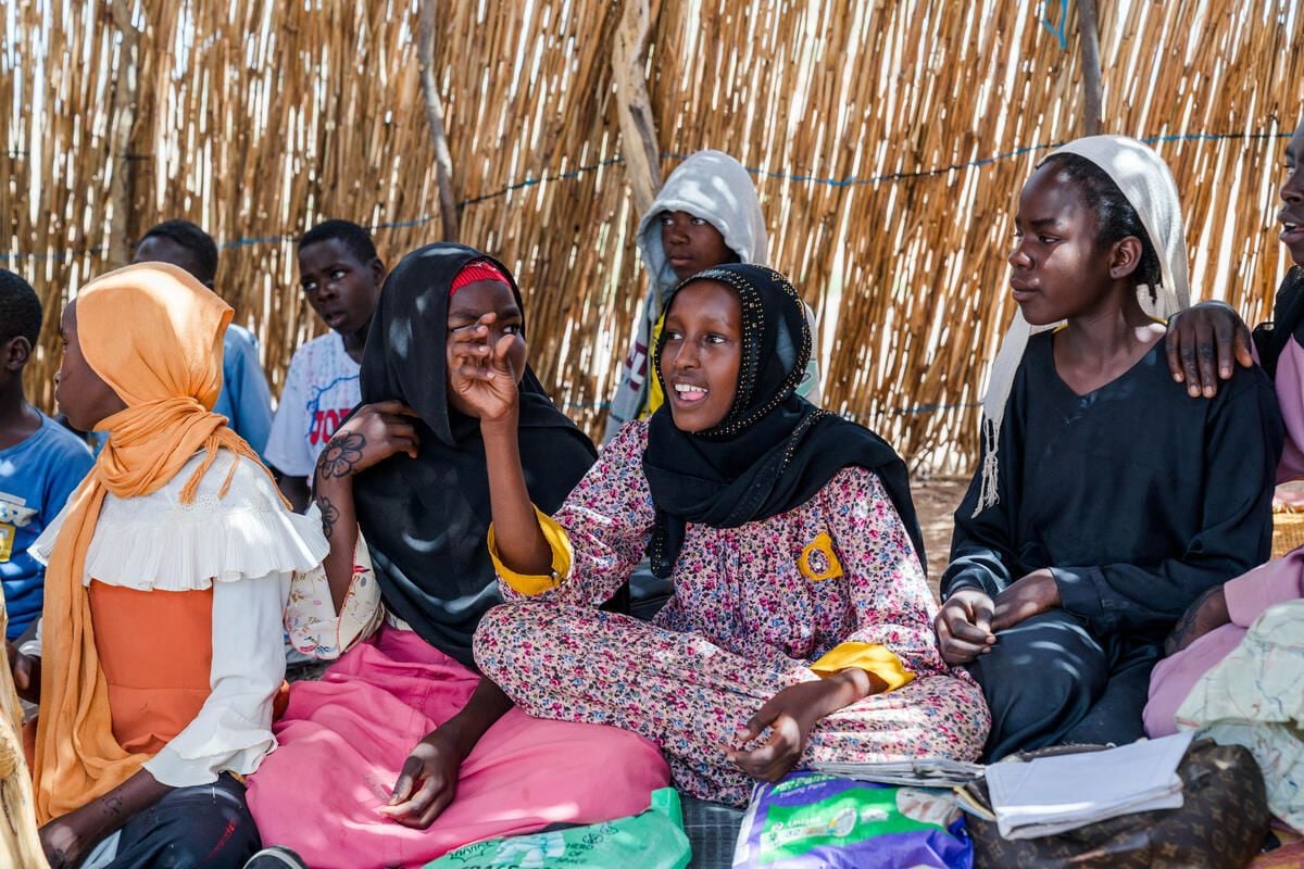 A group of students sit on the floor inside a thatch classroom; one girl in a floral dress speaks with her hand raised.