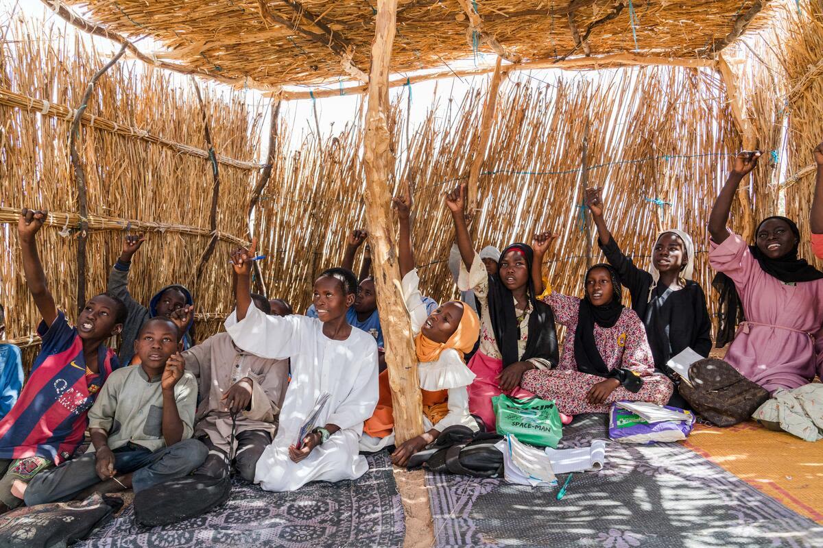 Children seated on mats inside a thatch-walled classroom raise their hands to answer a question during an outdoor lesson.