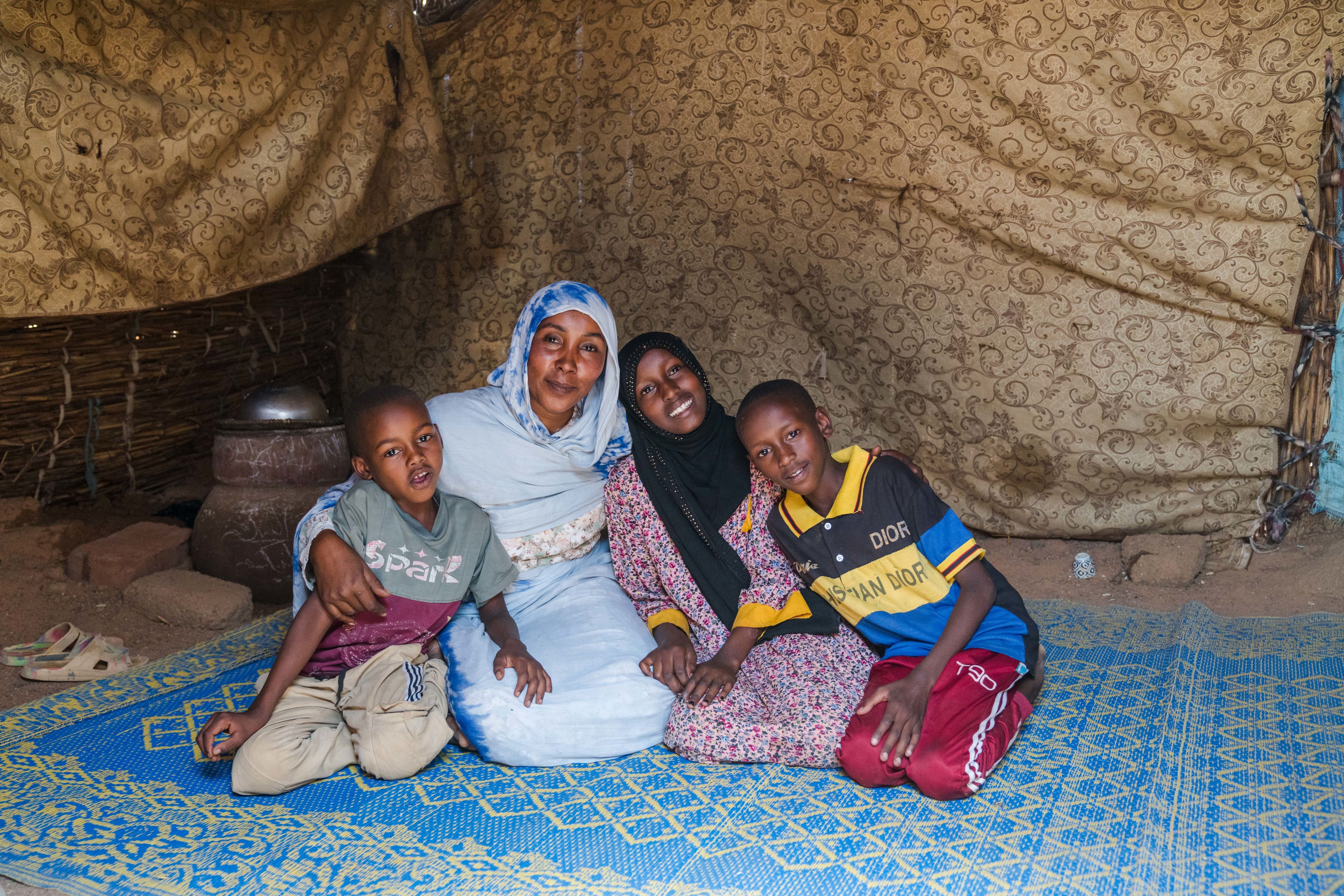 A woman sits on a blue mat inside a shelter with three smiling children, all leaning in close and posing for the camera.