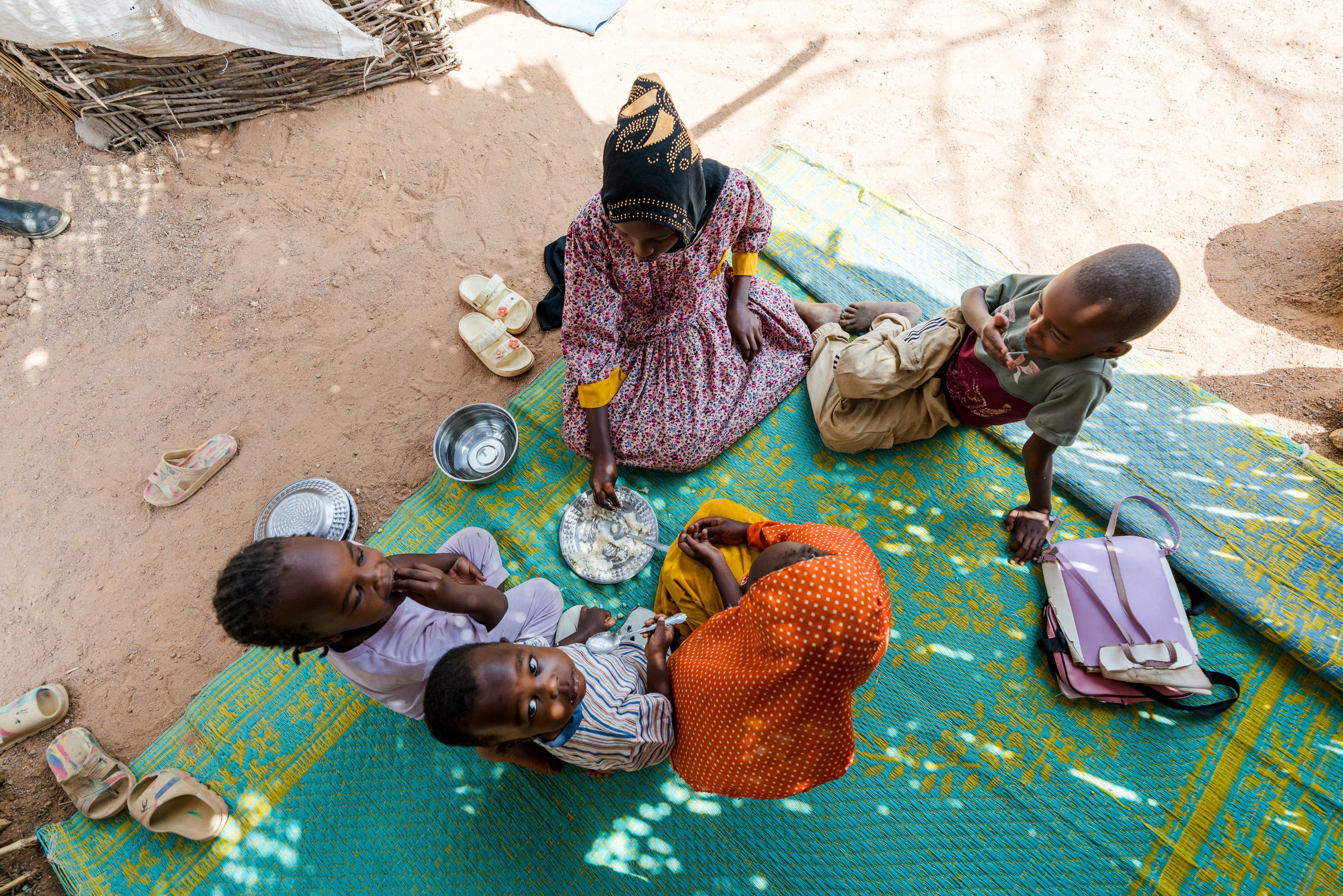 Four children sit on a woven mat outdoors, gathered around a shared plate of food, with sandals and dishes nearby.