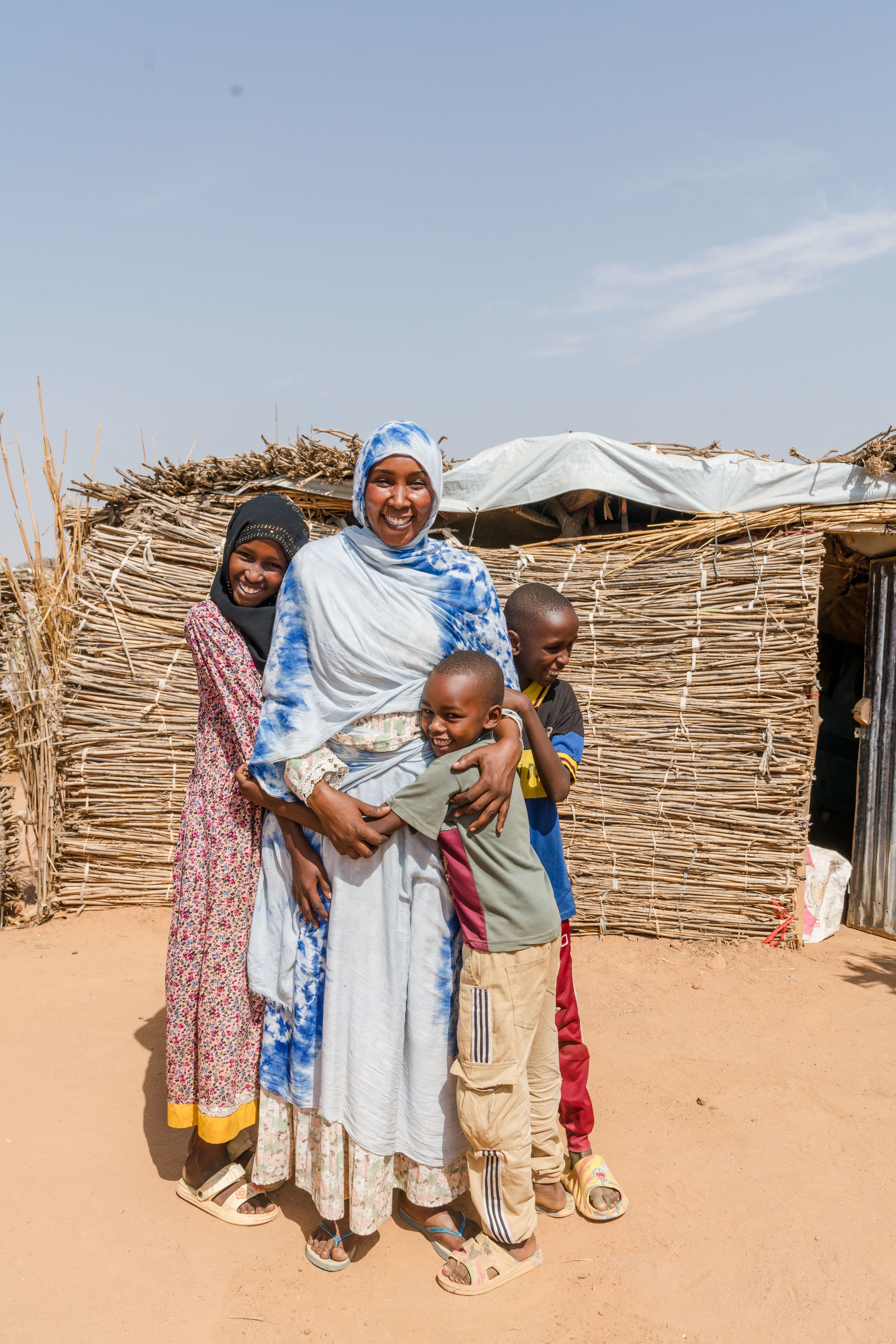 A smiling woman stands outside a shelter made of sticks and thatch, hugging three children closely.