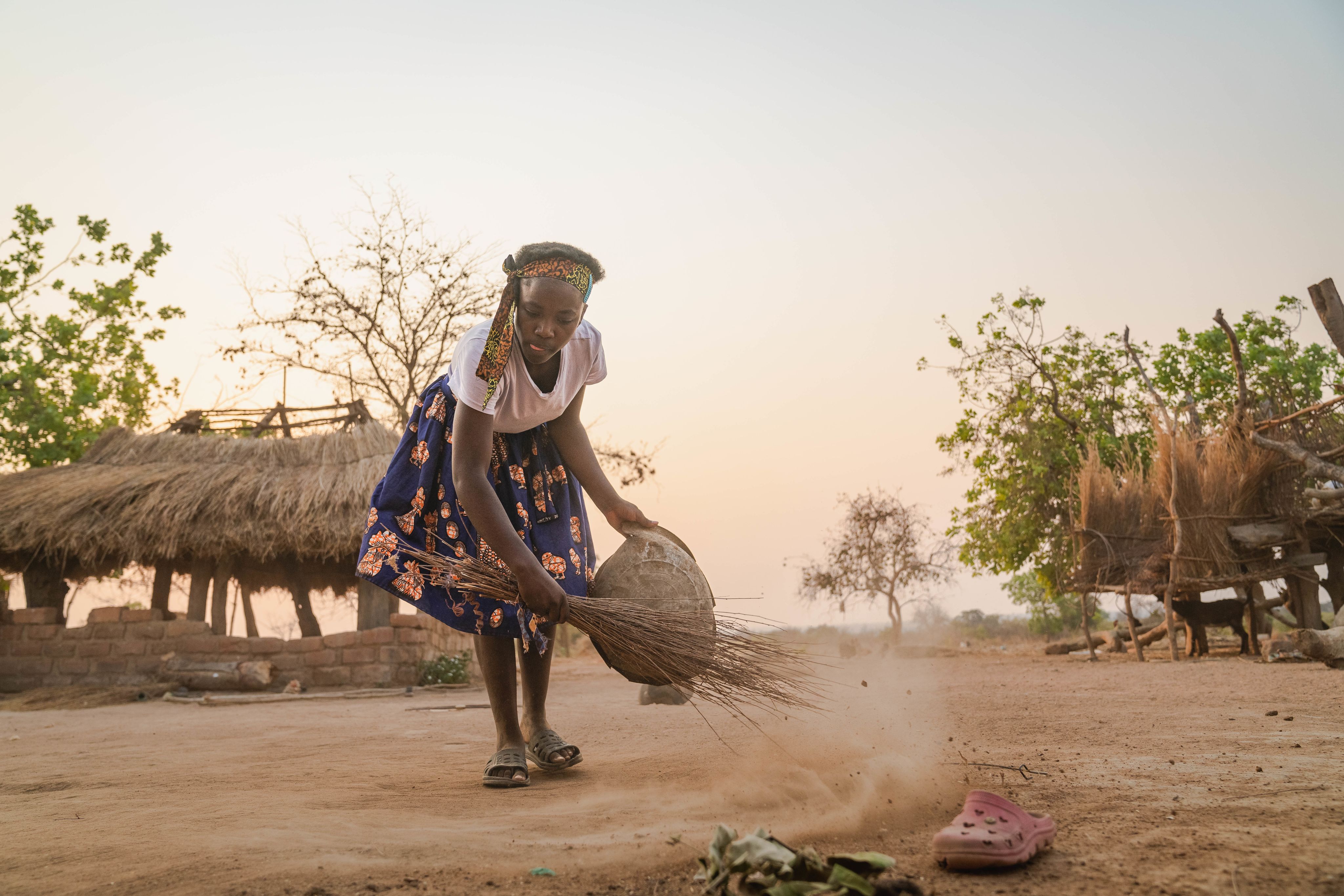 A young girl in a headband sweeps the dusty ground with a broom in a village at sunset.