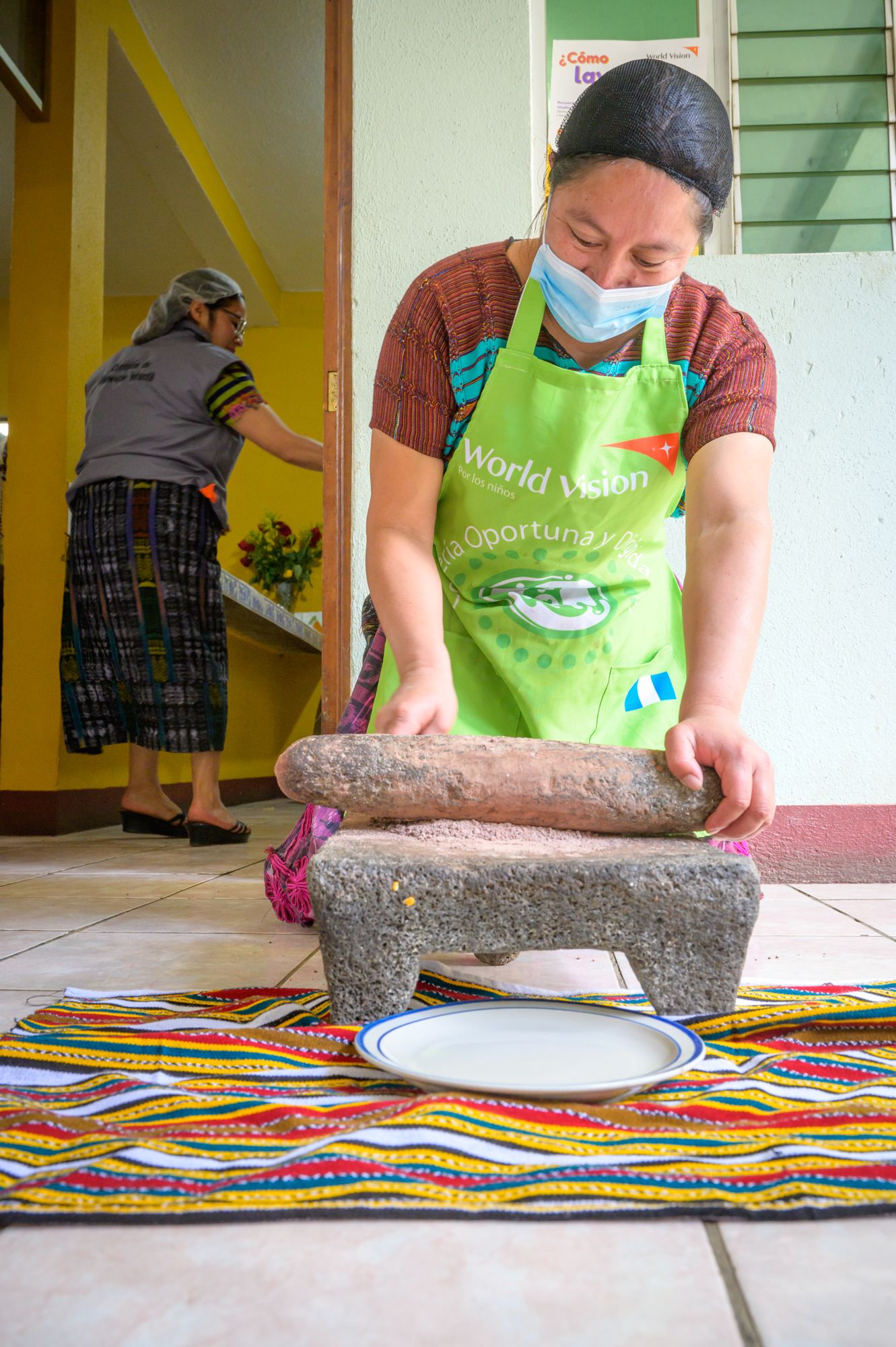 A woman wearing a mask grinds chocolate into a fine powder using a rolling pin on a stone.