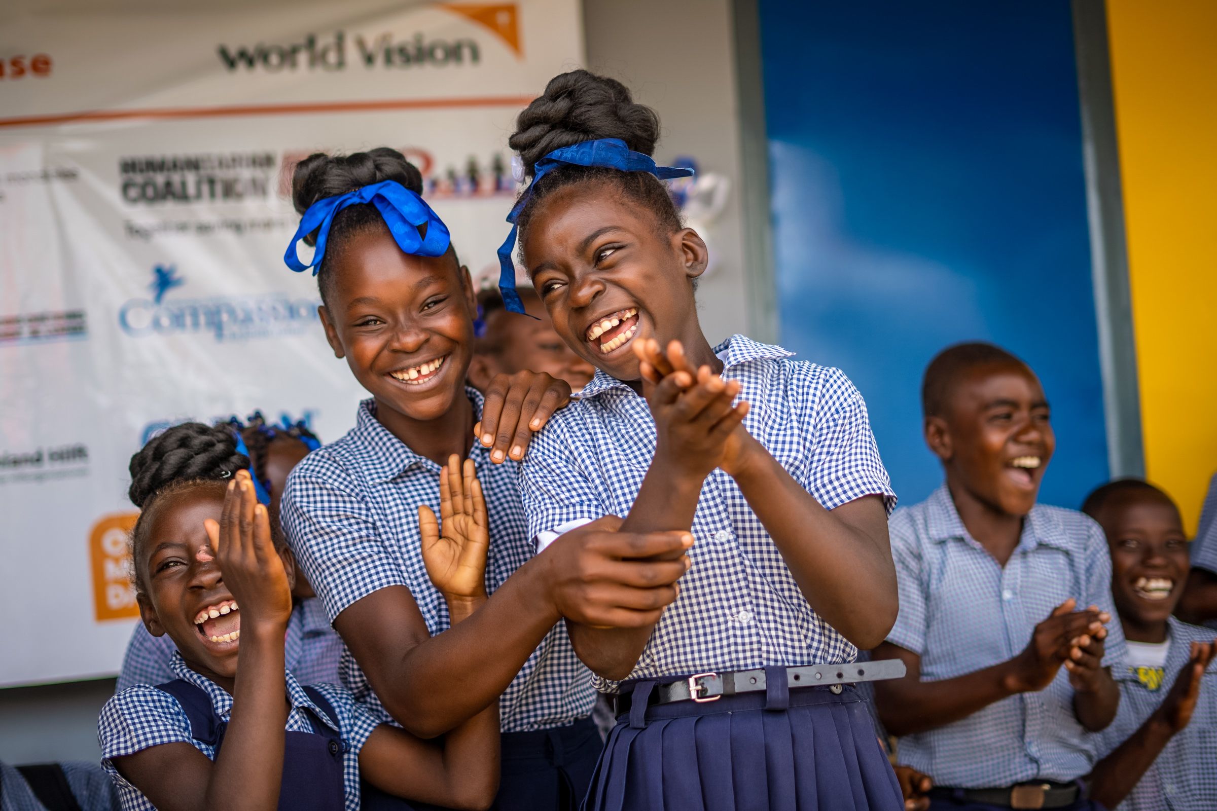 Children in school uniforms clap and laugh in celebration. In the background is a banner with logos, including World Vision’s.