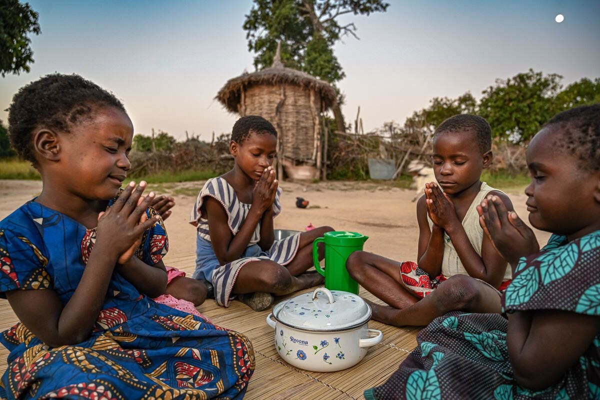 In Zambia, four children sit around a covered pot, praying. In the background the moon rises over a thatched hut. 