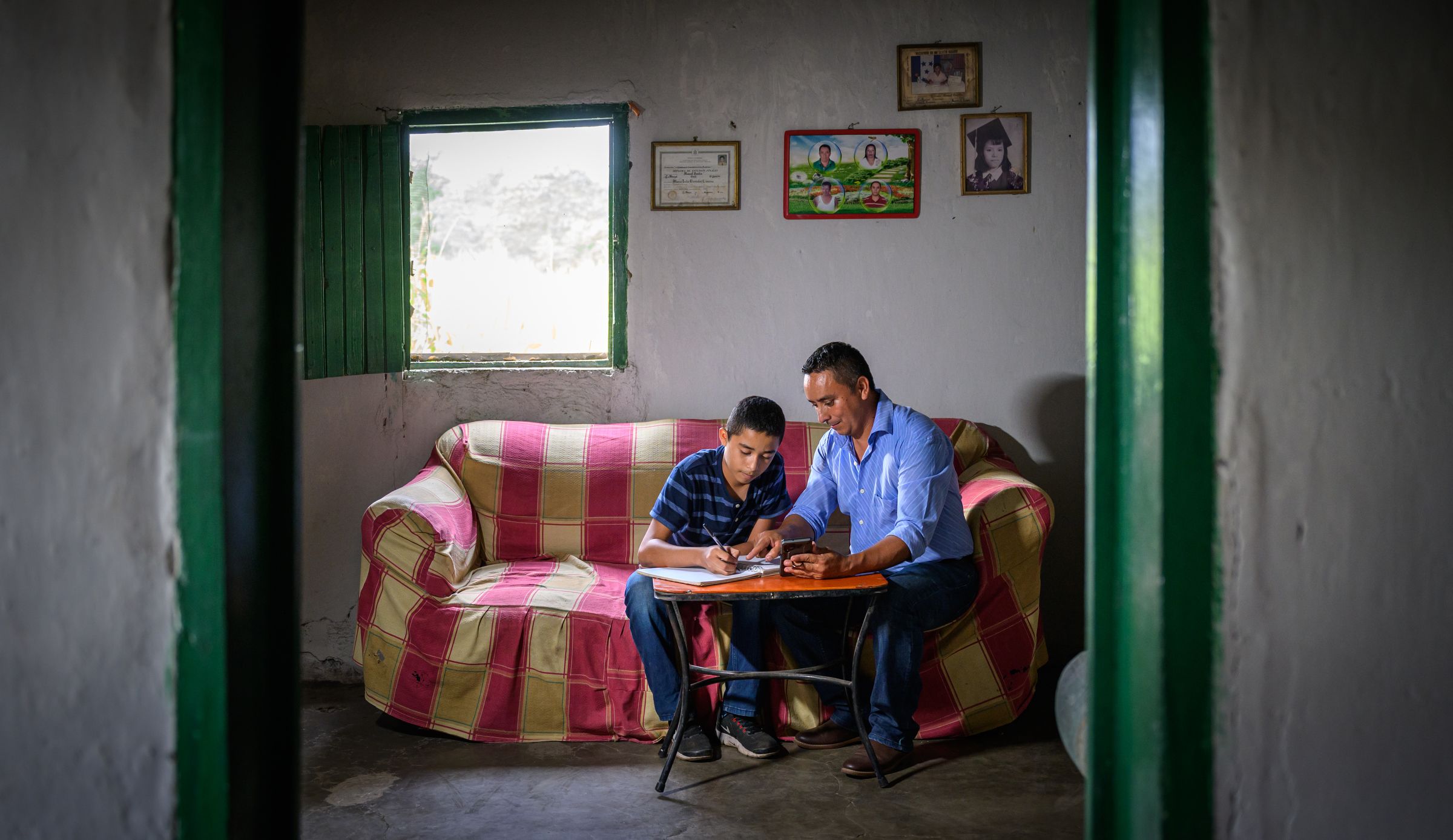Framed by an open green door and sitting on a couch, a father helps his son do schoolwork.