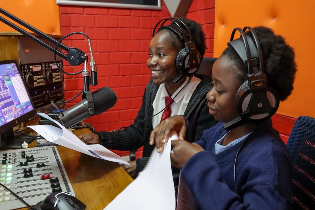 Two Zambian girls wear headphones in a sound booth. One holds a piece of paper while the other speaks into a microphone. 