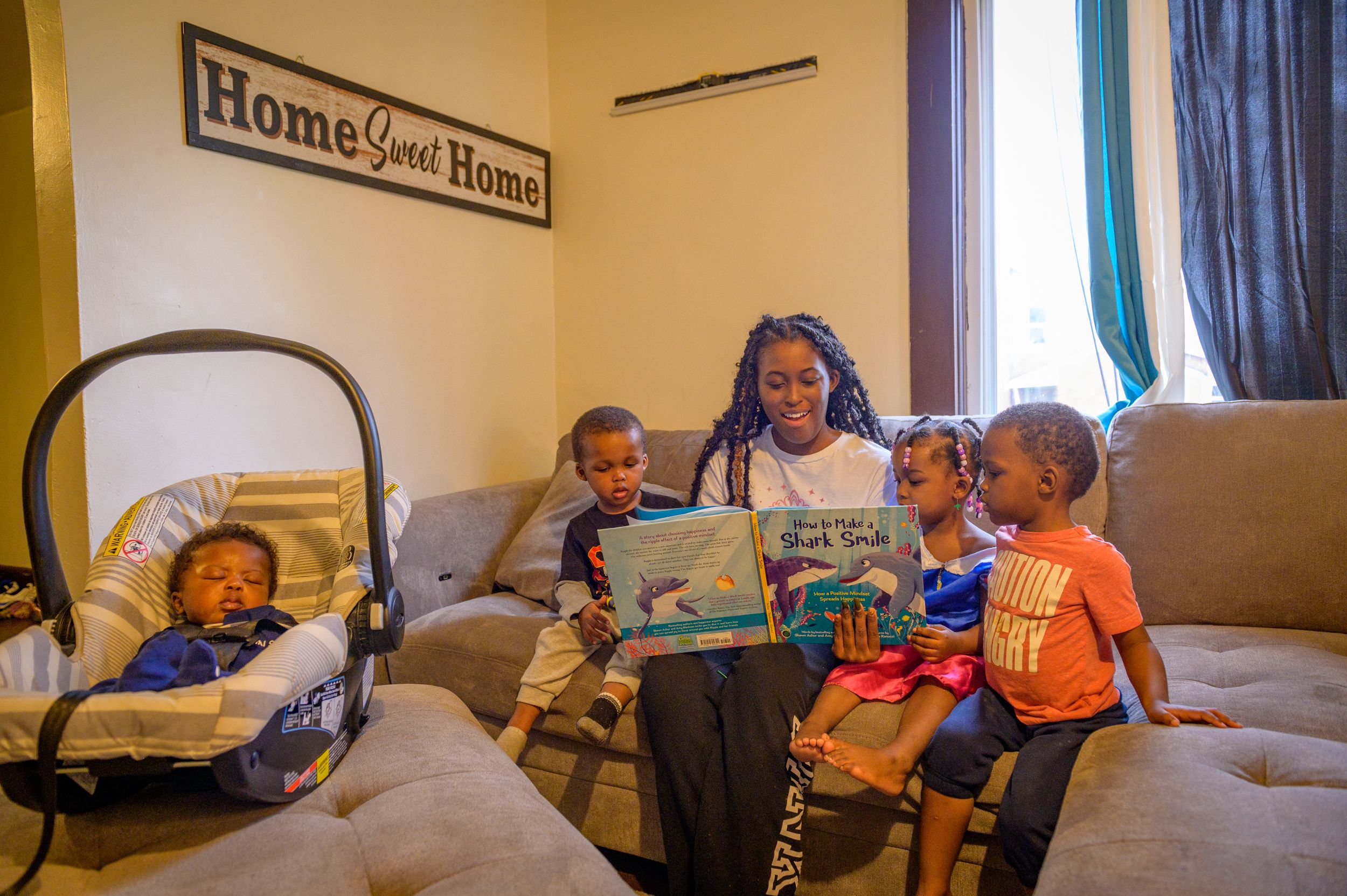 Jayla reads to her children, including a baby in a carrier, on the new sofa. Above them hangs a sign: Home Sweet Home.