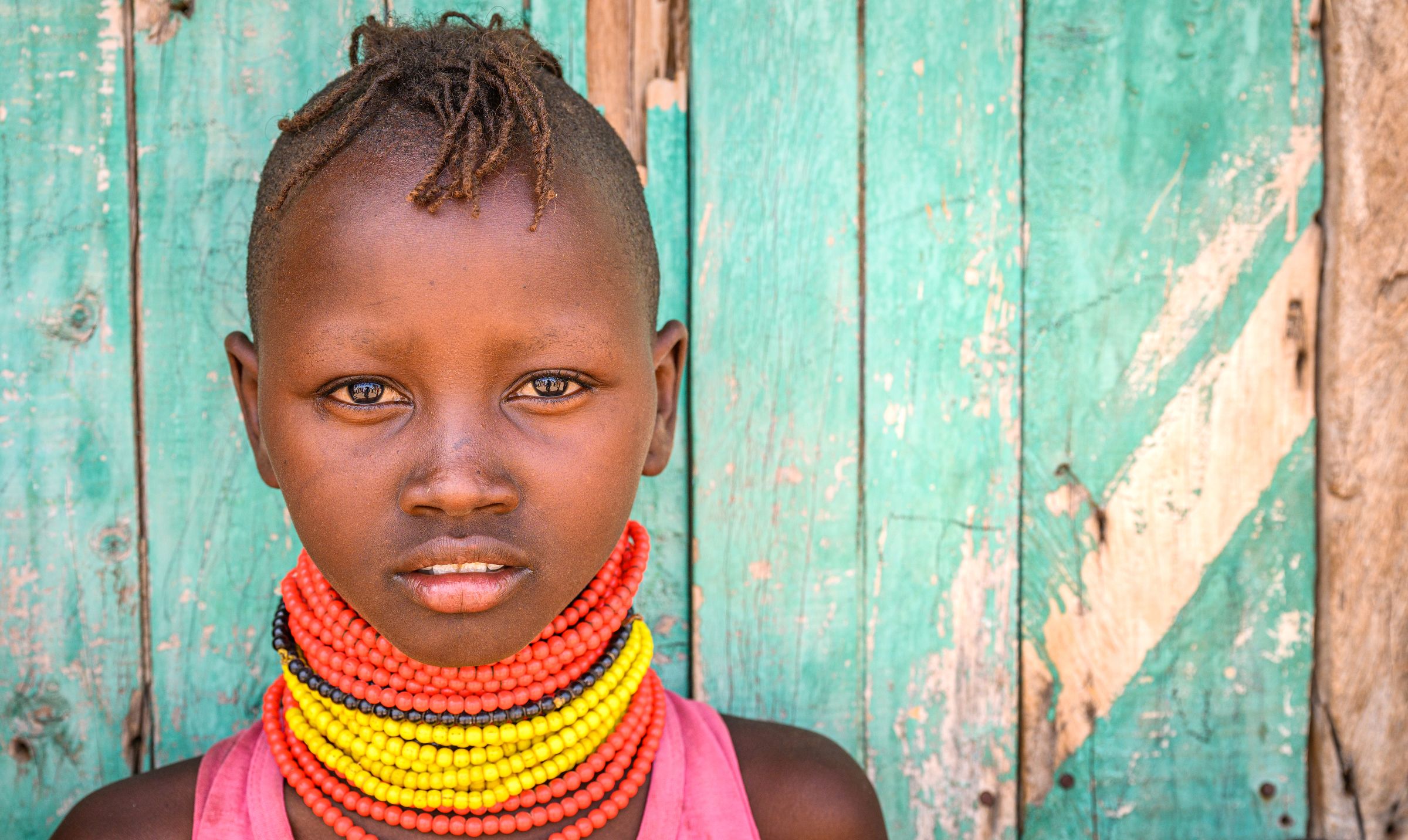 A girl in Turkana, Kenya, looks into the camera, red and yellow beads on her neck contrasting with a turquoise wall backdrop.