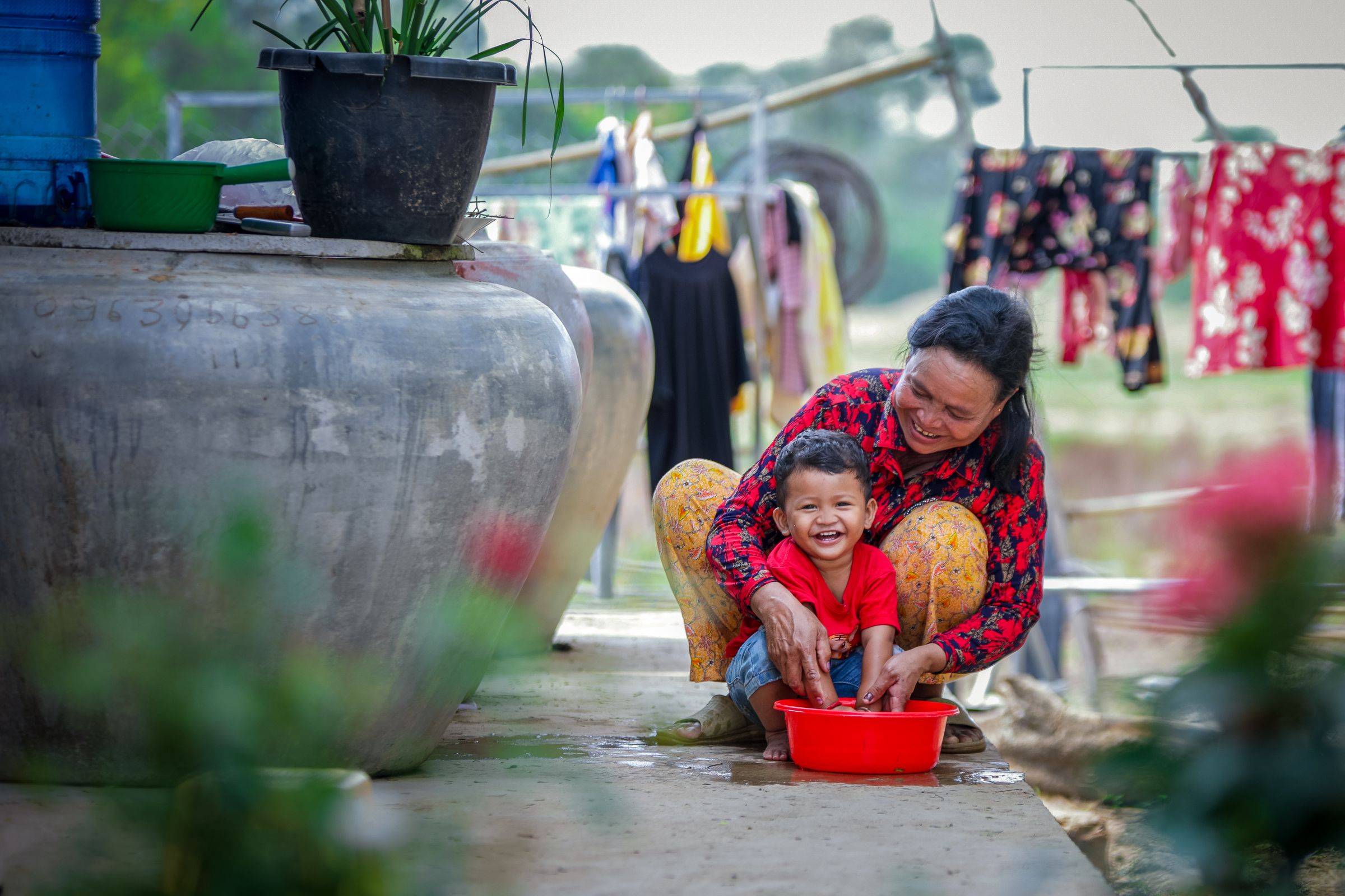 A grandma squats behind her smiling grandson, helping him wash his hands in a basin. To the left are giant water storage pots.