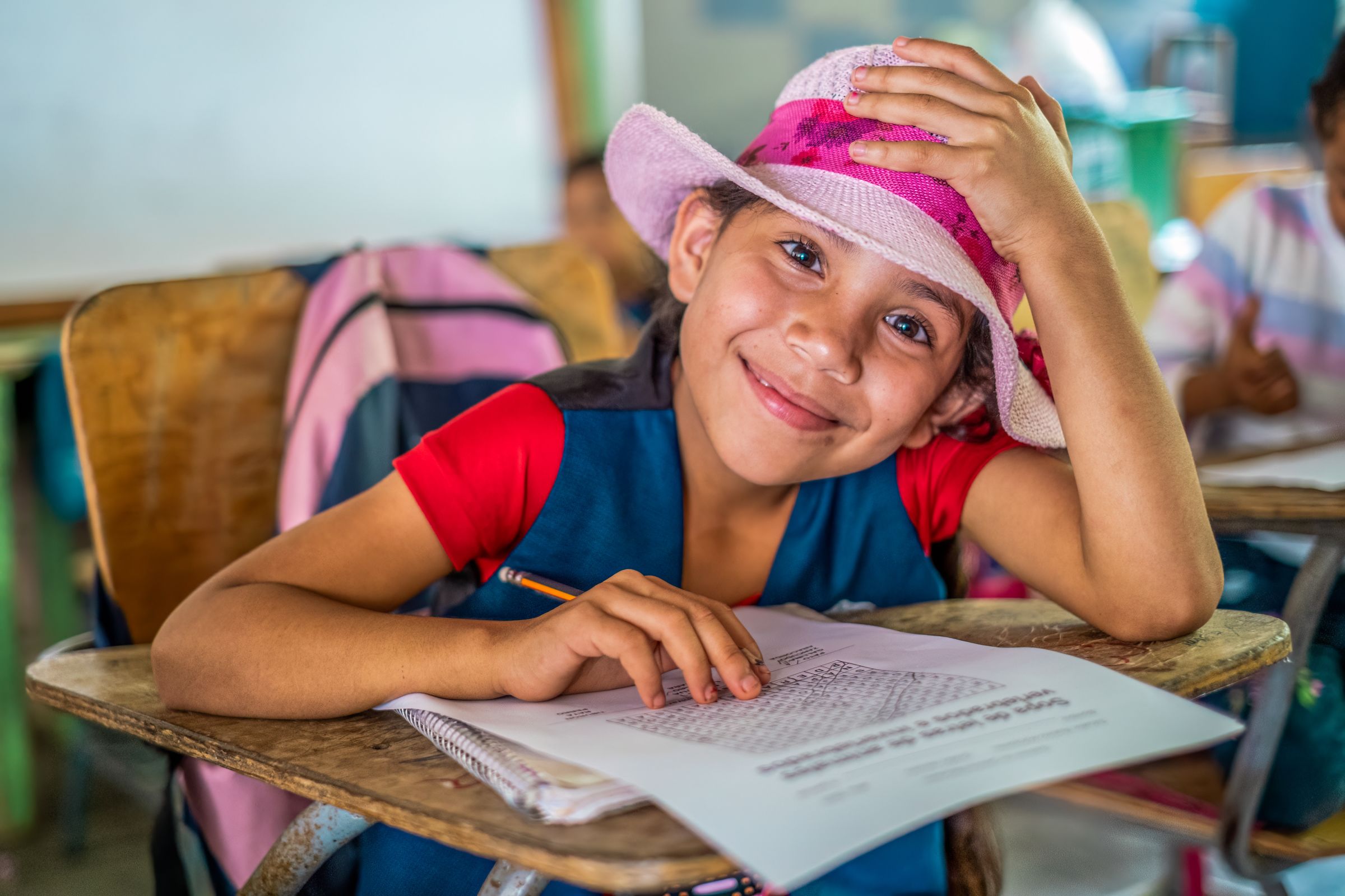 A young girls smiles at a classroom desk with her hand rested on her pink cowboy hat.