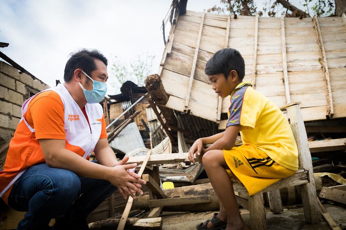 A World Vision staff member, wearing a mask, talks with a boy in yellow who is sitting in among the rubble of his home.