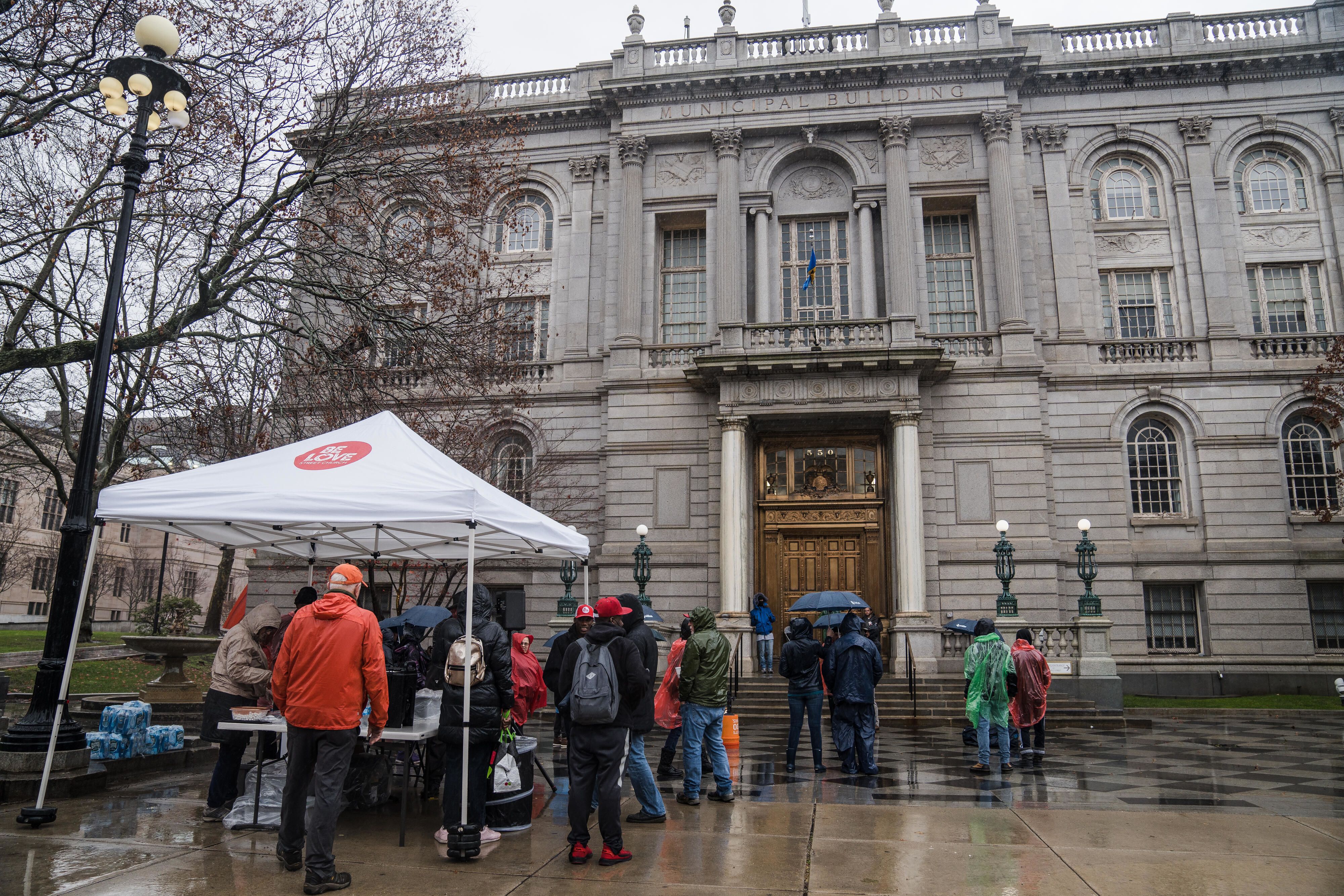 On a rainy day, people gather in a plaza in front of a historic-looking building. Under a tent, people serve hot drinks.