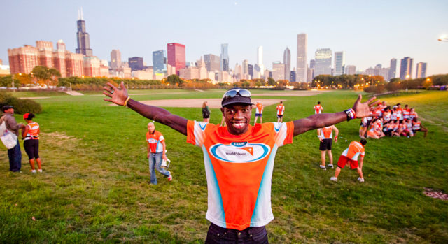 A man smiles with his arms stretched wide. He and others in the background wear Team World Vision shirts. The Chicago skyline is seen in the background.