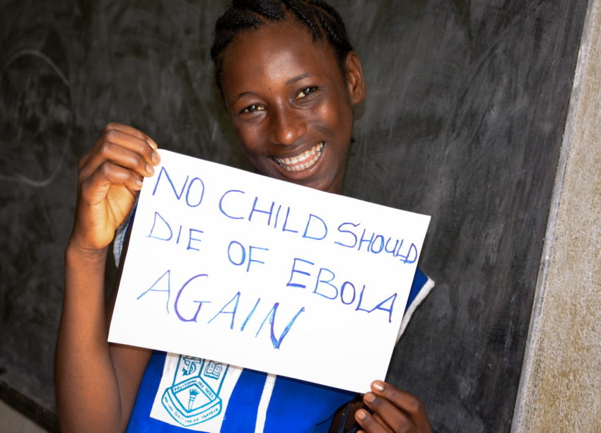 A Sierra Leonean teen smiles as she holds a sign that reads, “No child should die of Ebola again.”