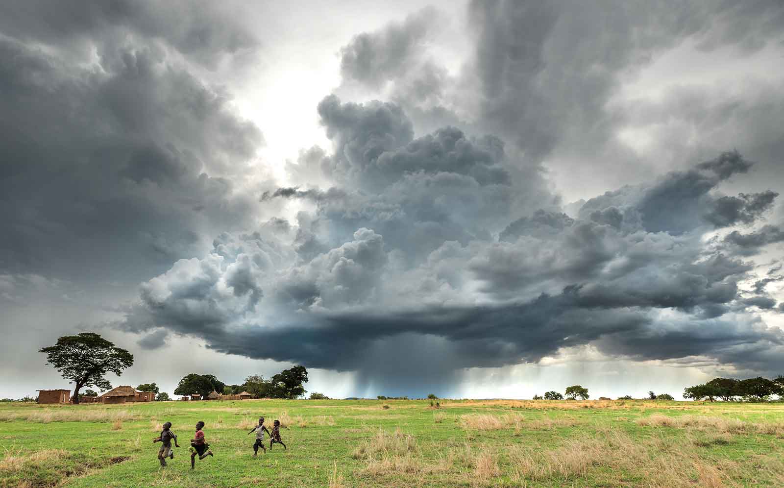 Children run across a field as stormy cumulonimbus clouds pour rain to a village in Magoye, Zambia.