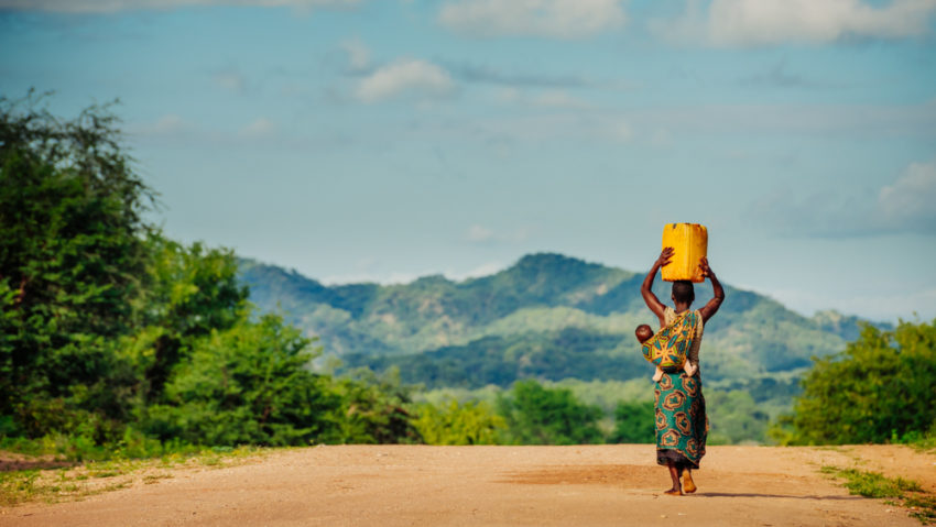 A woman in Sinazongwe Zambia, carries a baby on her back and container of water from a World Vision borehole on her head. Carrying water long distances can have lasting effects on one’s health, some research shows. (©2016 World Vision/photo by Jon Warren)