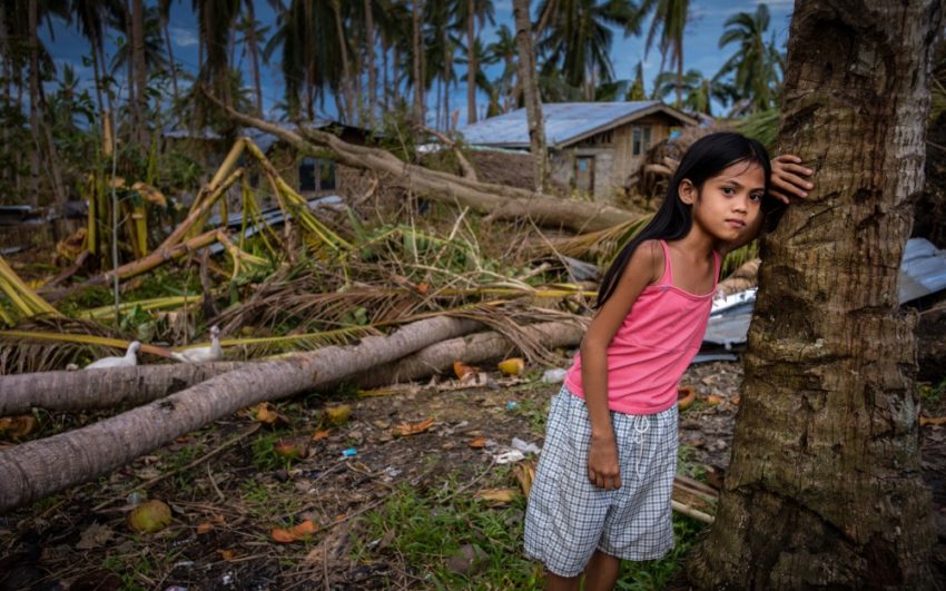 Girl standing against a coconut tree with destroyed house in background in the Philippines. Pray for the Asia-Pacific Islands.