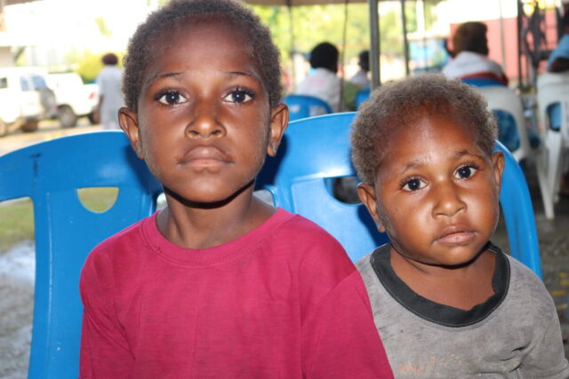 Two young siblings wait, seated on blue plastic chairs, eyes meeting the camera.