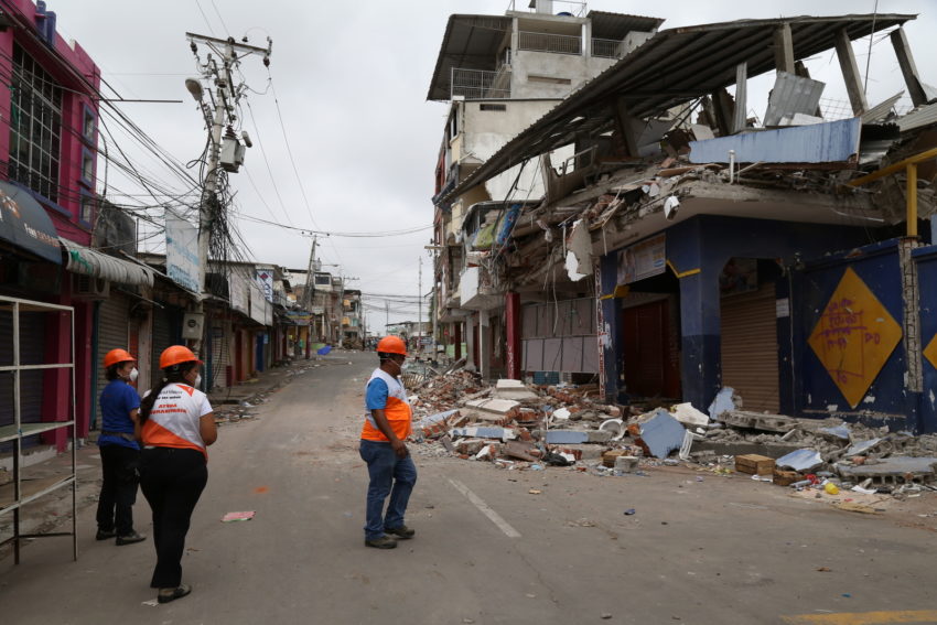 Three people wearing World Vision shirts and hard hats look at a severely damaged building.