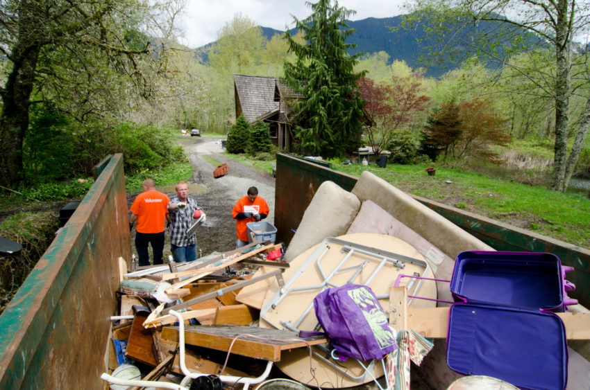 Three men, two wearing bright orange T-shirts with the World Vision logo, help throw debris and broken furniture into a huge dumpster.