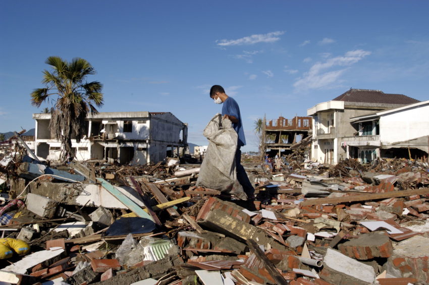 A man carries a large canvas bag with items salvaged from the rubble. A blue sky contrasts the devastation around him.