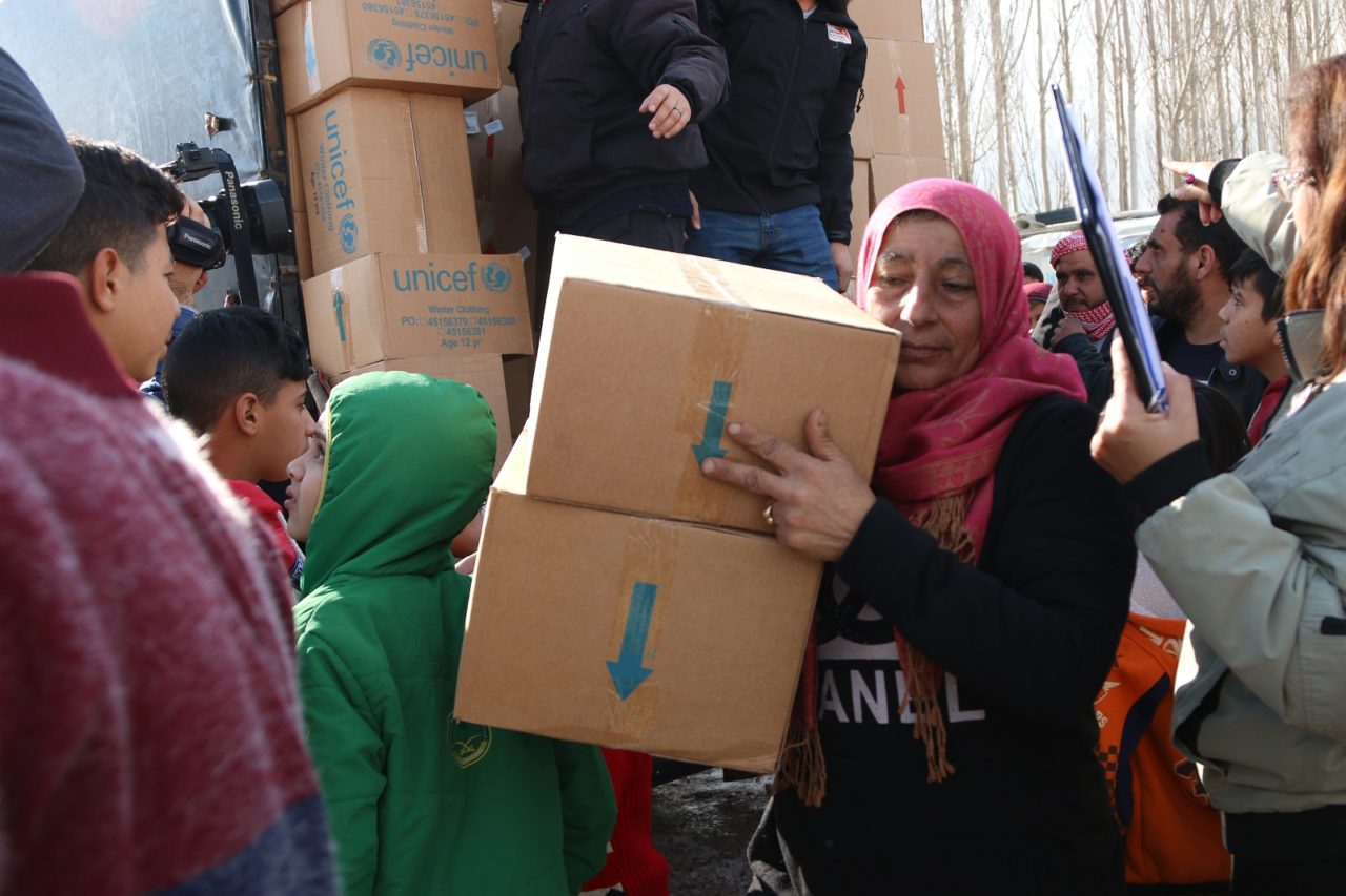 A woman carries boxes toward the camera amid a group of people around boxes with the UNICEF logo.