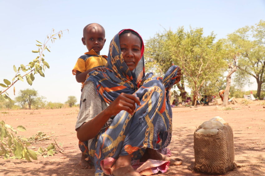 A woman looks at the camera, with a child standing behind her in the shade on dry, sunlit ground, near their water containers.