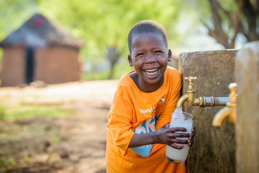Happy Cheru, wearing a World Vision Global 6K tee-shirt, fills her glass at the water point at her home, and takes a drink of clean water. Cheru, 6, benefits from a 16-kilometer, gravity-fed World Vision pipeline that brings clean water to her community. Along with other children, she used to walk 6 kilometers daily to carry water to school and home. Now the pipeline brings water to both places.