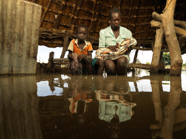 A young boy and a woman holding a baby sit on a bench under a hut while floodwater reaches their shins.