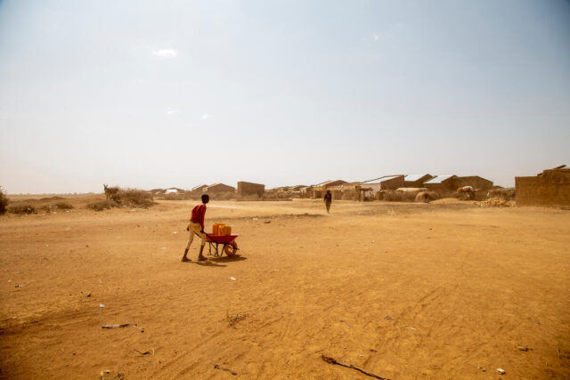 A boy pushes a red wheelbarrow containing two yellow jerrycans across a flat, sandy terrain.