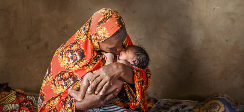 A mother in an orange head covering kisses the nose of her newborn baby as she holds him close in her hands.