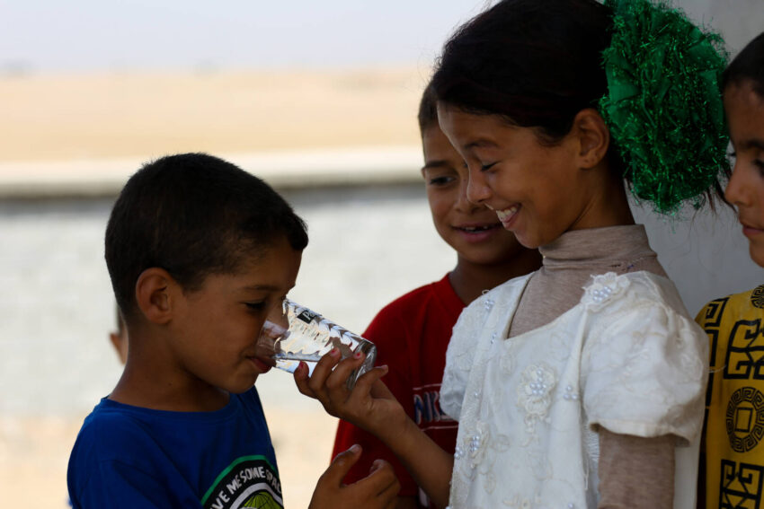 A smiling girl holds a clear glass of water as another child drinks from it.