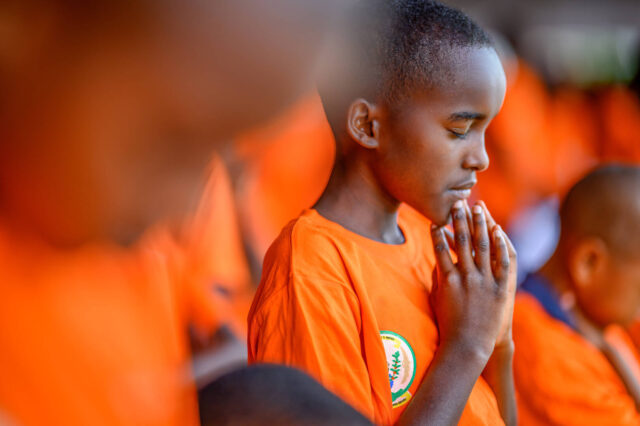 Portrait of a praying child in an orange shirt surrounded by other children in orange shirts in soft focus.