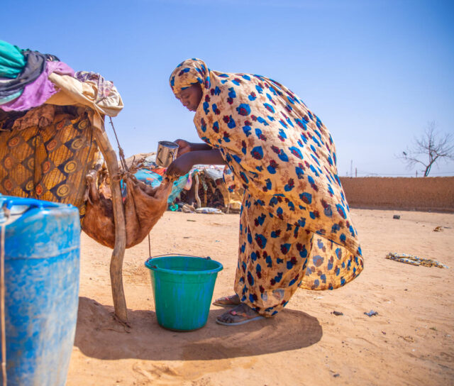 Woman in a colorful headscarf and dress bends on dry ground to fill a goatskin water container under a bright blue sky.