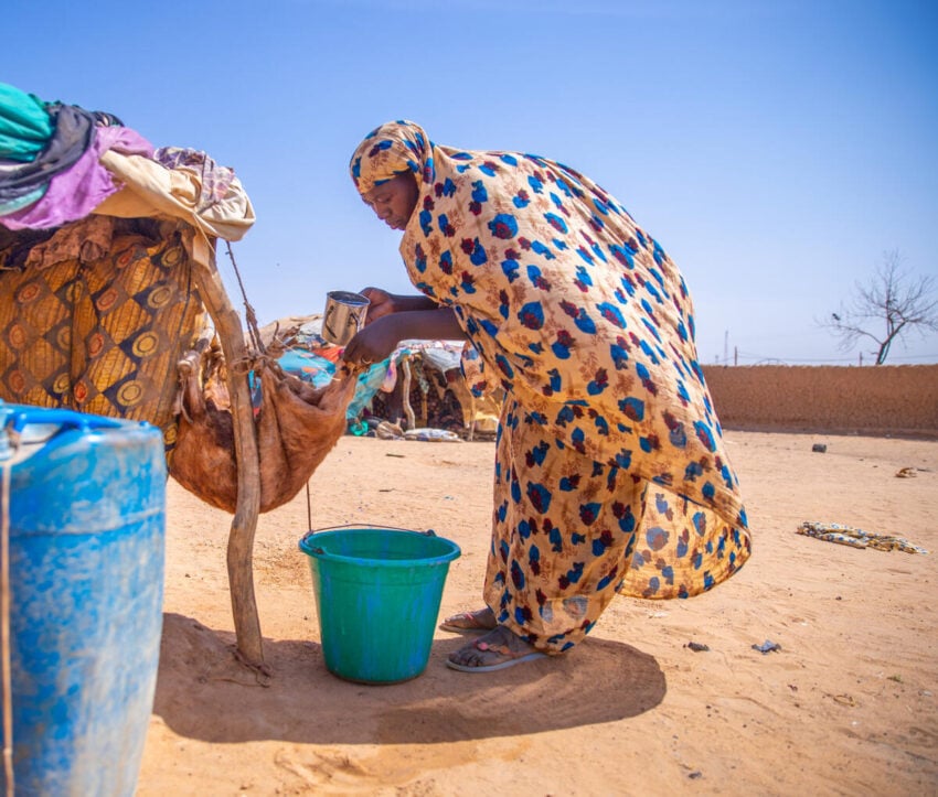 Woman in a colorful headscarf and dress bends on dry ground to fill a goatskin water container under a bright blue sky.