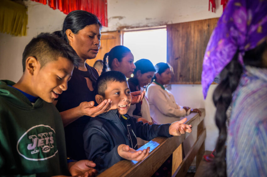 A Honduran mother and two boys pray with uplifted hands standing in a church pew.