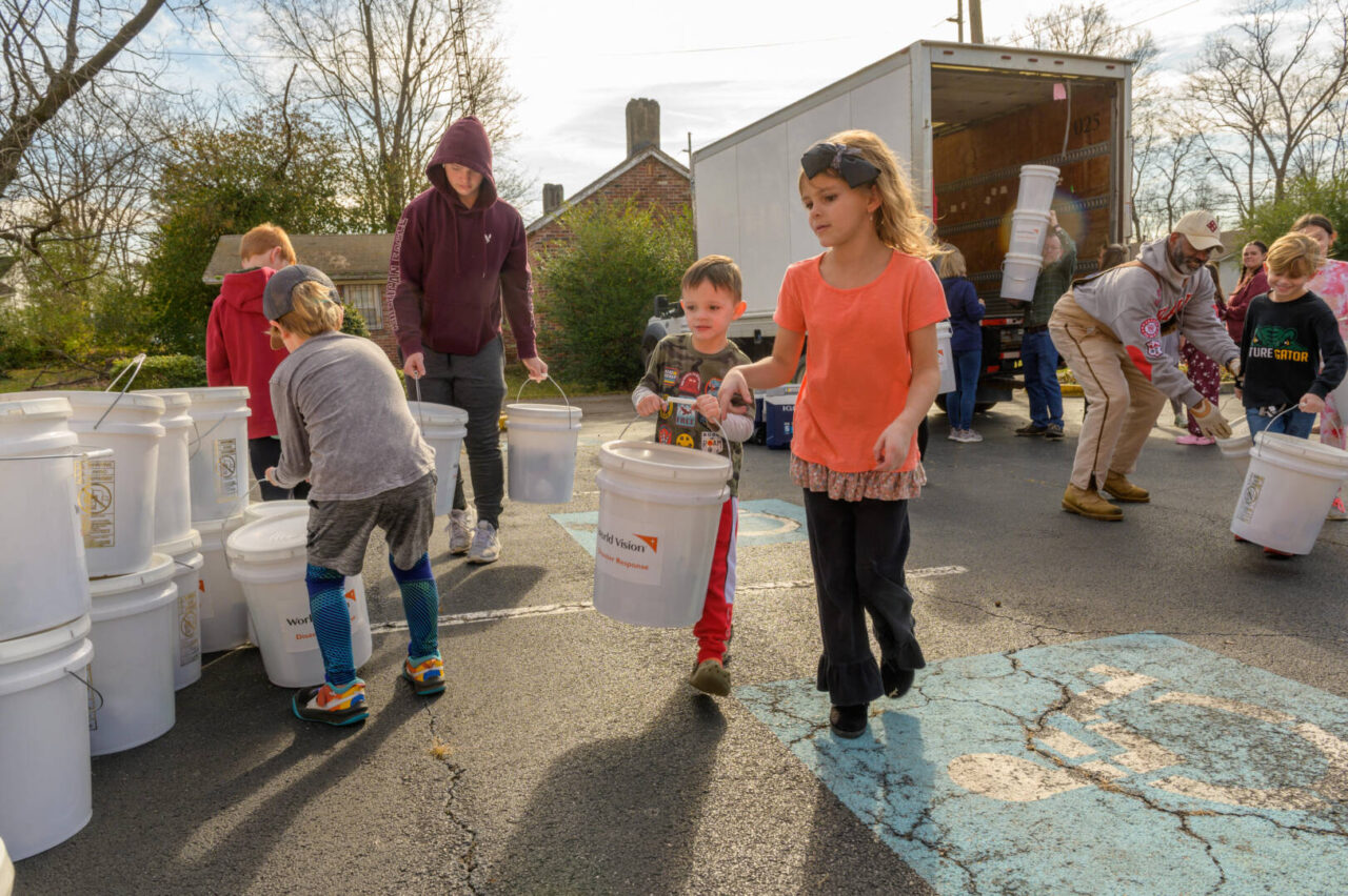 Volunteers unload buckets of relief supplies from a truck. Children carrying a bucket together are in the foreground.