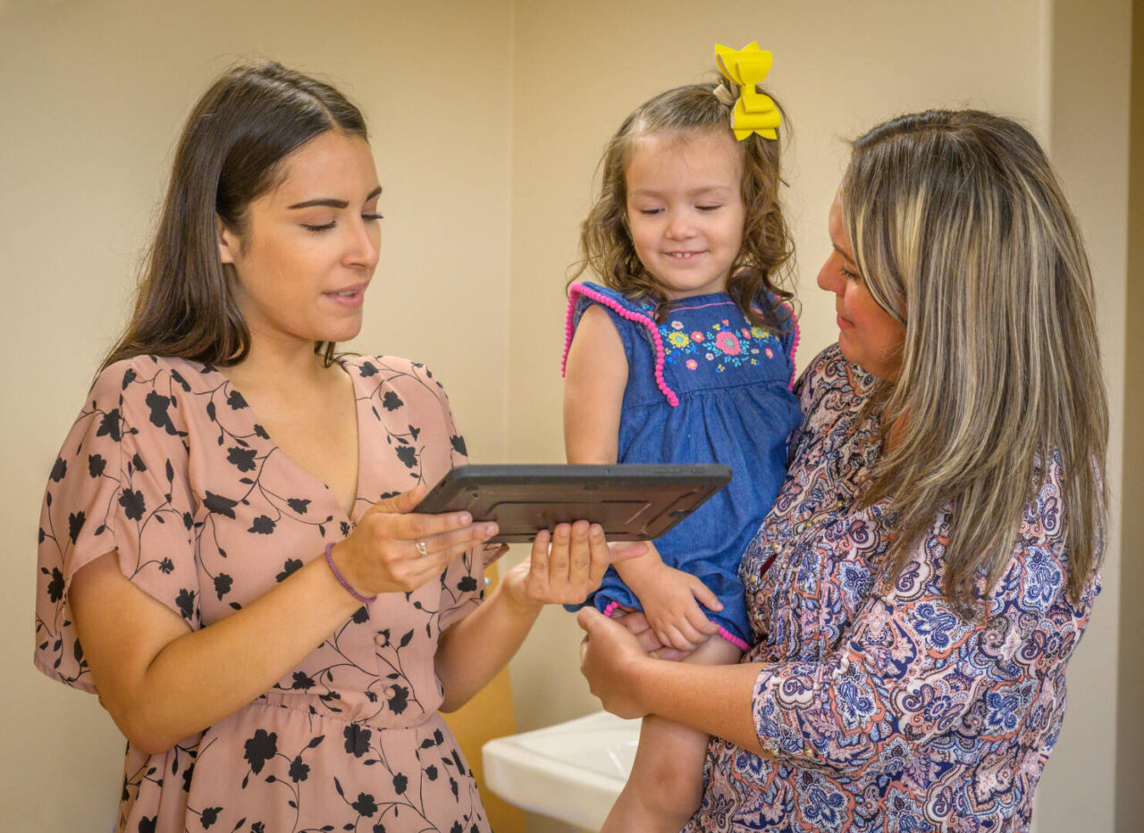 Two women hold up a tablet for a little girl to read from.