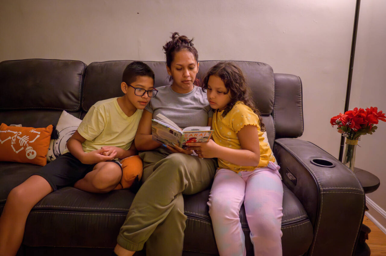 A woman sits between her two children on a couch, reading a book to them.
