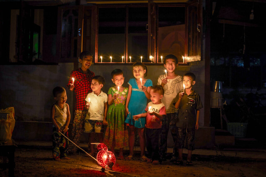 At nighttime, a small group of children in Myanmar smile and laugh as they hold up candle sticks, the light of the flames glowing on their faces.