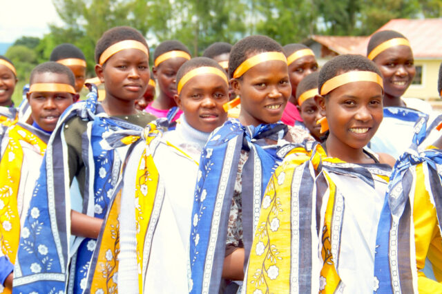 A group of Kenyan girls, each wearing a blue, yellow, and white patterned fabric tied at their shoulders and yellow headbands.