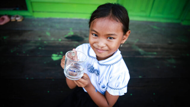 A girl wearing a school uniform looks at the camera while holding a cup of water with P&G and World Vision logos on it.