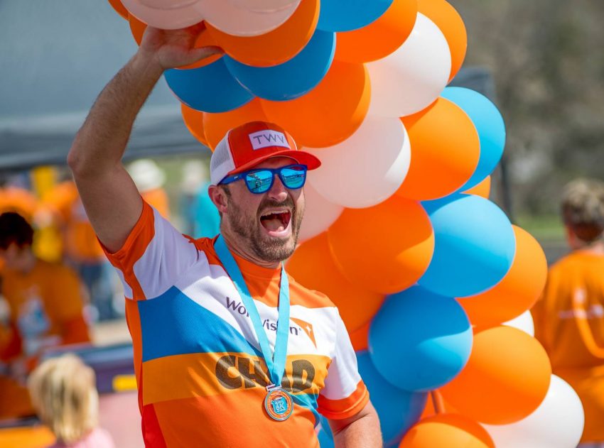 A man in orange and blue shirt stands with one arm raised up to orange, white, and blue balloons.